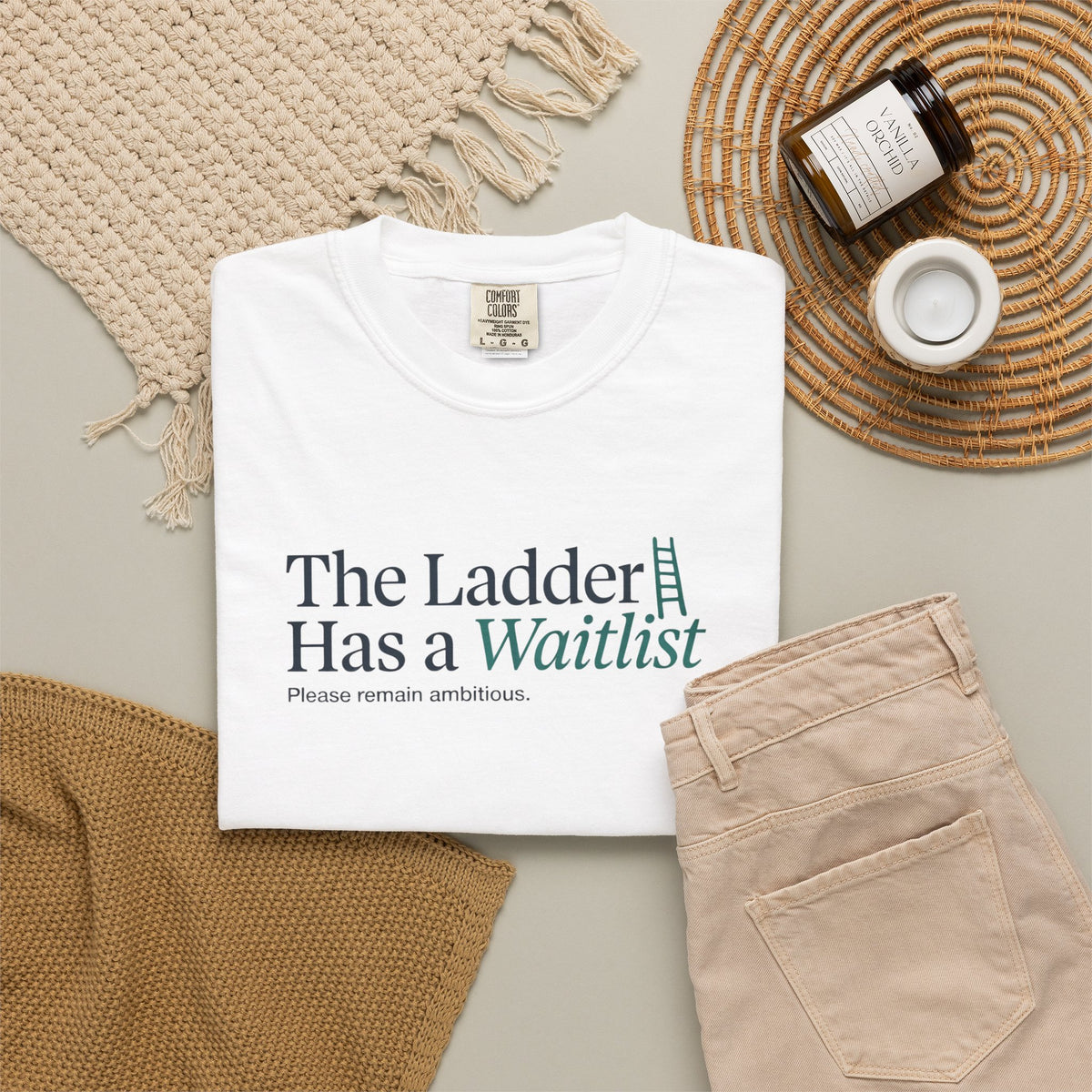 "The ladder has a waitlist" garment-dyed unisex tee in White, folded flat lay with a knit throw and a candle — Givvo