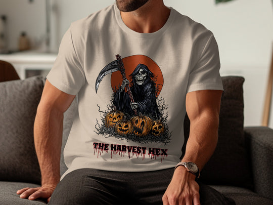 Person wearing a sand color t-shirt with a graphic design of the Grim Reaper and pumpkins, labeled 'The Harvest Hex', sitting on a couch.