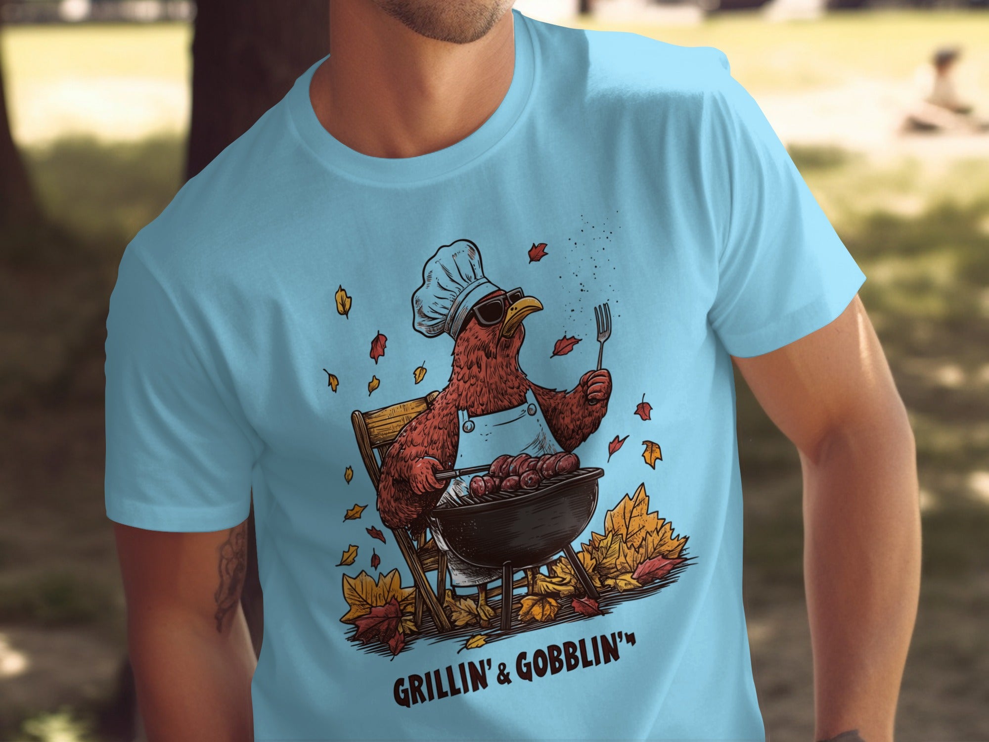 Person wearing a sky blue t-shirt with a graphic of a chicken cooking on a grill and the text 'Grillin' & Gobblin'.