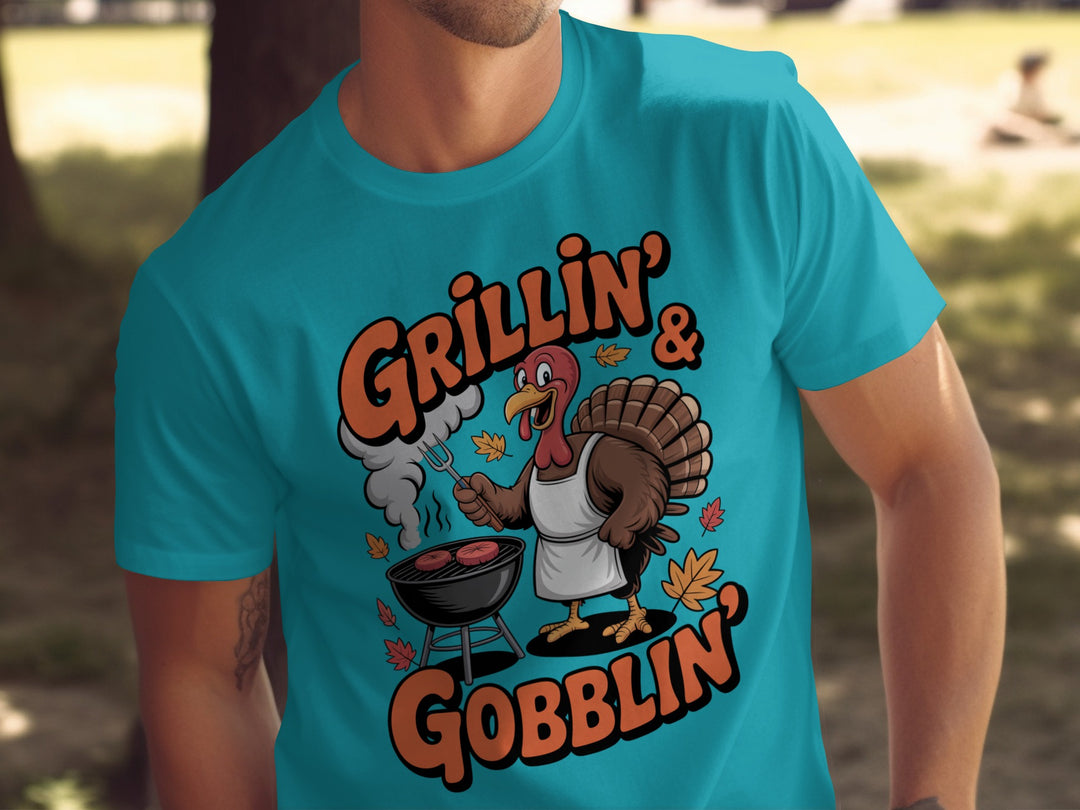 Person wearing a turquoise t-shirt with a graphic of a turkey grilling and text 'Grillin' & Gobblin'.