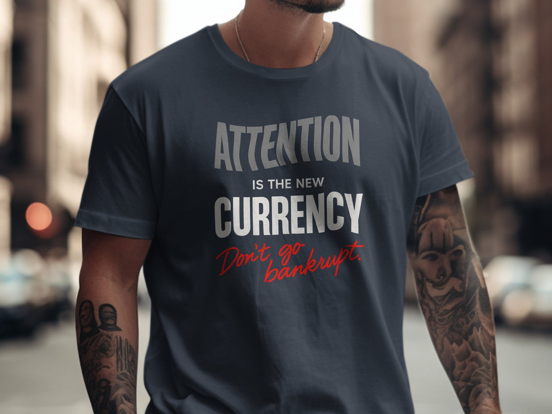 Person wearing a navy blue t-shirt with text 'Attention is the new currency. Don't go bankrupt.' on a city street.